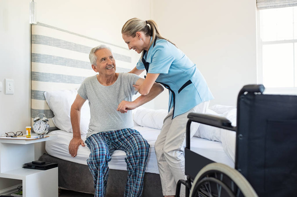Smiling nurse assisting senior man to get up from bed. Caring nurse supporting patient while getting up from bed and move towards wheelchair at home. Helping elderly disabled man standing up in his bedroom. - Photo, image