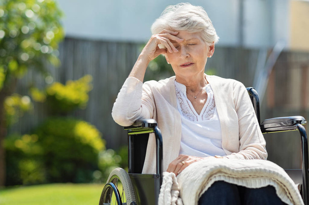 Smutny starszy kobieta siedzi na wózku inwalidzkim na zewnątrz domu opieki. Przygnębiony niepełnosprawnych Pani z straszny ból głowy. Samotność i porzucenia koncepcji. - Zdjęcie, obraz