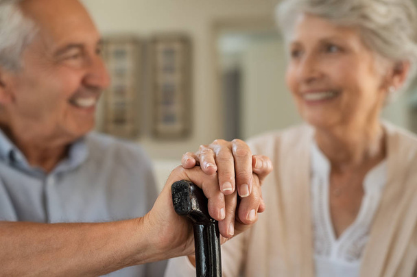 Closeup of senior couple hands holding walking stick. Old husband and wife holding cane handle while looking at each other. Happy retired couple joining hands with love at home. - Foto, Bild