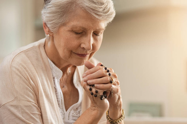 Senior woman praying while holding christian symbol of crucifix. Old woman praying to god with hope and closed eyes. Elderly believer make a prayer with faith holding rosary in hands.  - Zdjęcie, obraz