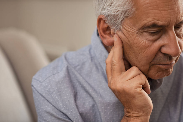 Visão lateral do homem idoso com sintoma de perda auditiva. Homem maduro sentado no sofá com os dedos perto da orelha sofrendo dor
. - Foto, Imagem
