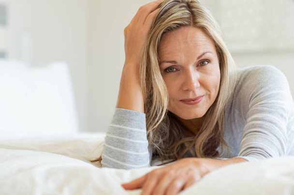 Thoughtful woman laying on bed in bedroom and looking at camera. Portrait of middle aged woman smiling. Portrait of a beautiful middle aged woman at home. - Foto, afbeelding