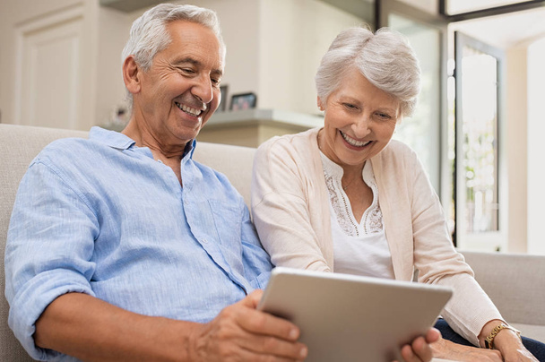 Senior couple using digital tablet sitting on couch at home. Happy old man surfing the net with his wife. Retired man and smiling elderly woman planning on summer vacation using tablet.  - Foto, afbeelding