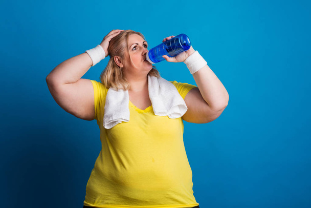 Portrait of a tired overweight woman in studio on a blue background, drinking water. - Foto, imagen
