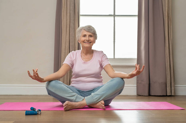 Senior smiling woman doing yoga in her living room. Elderly relaxed woman sitting in lotus pose and meditating while practicing yoga at home. Old grandmother sitting on yoga mat smiling. - Photo, Image