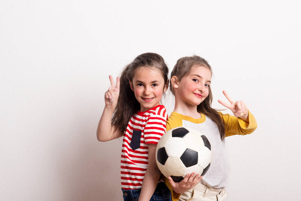 Маленькие девочки с футбольным мячом стоят в студии, показывая знак победы
. - Фото, изображение