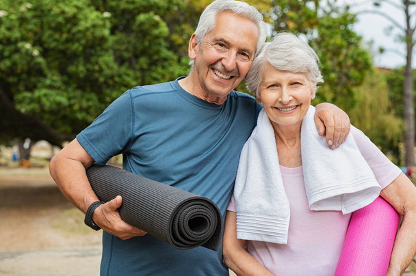 Lovely senior man and old woman holding yoga mat for exercising outdoor. Aged husband and wife ready for yoga session at park. Active healthy retired couple looking at camera before a postural session. - Foto, afbeelding