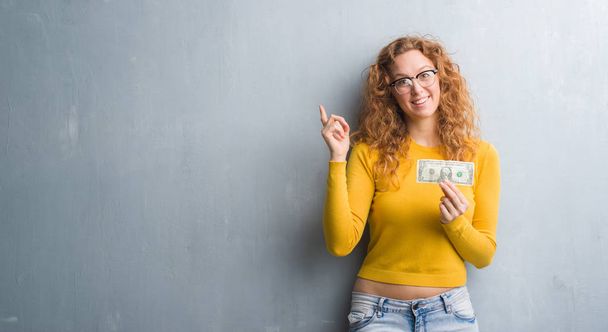 Young redhead woman over grey grunge wall holding a dollar very happy pointing with hand and finger to the side - 写真・画像