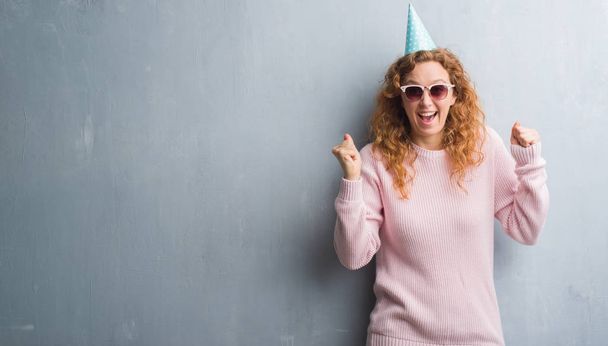 Mladá zrzka žena přes zeď šedá grunge nosí narozeniny cap křičí hrdá a slaví vítězství a úspěch, velmi vzrušený, fandění emoce - Fotografie, Obrázek