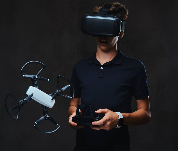 Der junge Teenager im schwarzen T-Shirt trägt eine Virtual-Reality-Brille und steuert den Quadrocopter mit der Fernbedienung. isoliert auf dunklem Hintergrund. - Foto, Bild