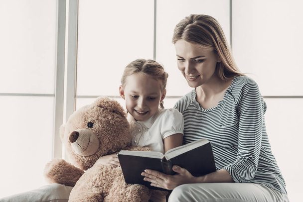 Joven madre leyendo el libro a la pequeña hija. Hermosa mujer sonriente leyendo a linda hija adorable sosteniendo oso de peluche. Mamá le lee a Child en casa. Concepto Familia y Maternidad
 - Foto, Imagen