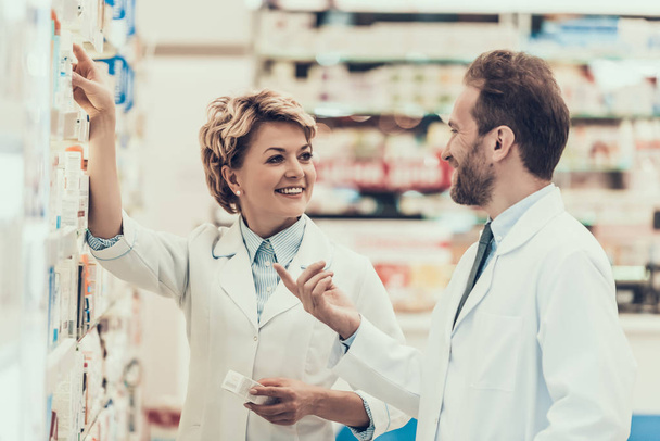 Female and Male Pharmacist Working in Drugstore. Two Pharmacist Standing next to Shelves with Drugs and checking Medicines Inventory in Pharmasy. Pharmacist Holding package of Medicine - Fotó, kép