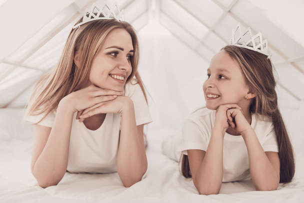 Young Mother and Little Doughter wearing Tiaras. Beautiful Smiling Woman spending time with Cute Adorable Daughter. Mom and Child under tent in Bedroom. Family and Motherhood Concept - Photo, image