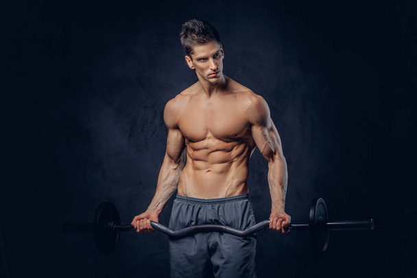 Handsome shirtless man with stylish hair and muscular ectomorph doing the exercises with the barbell on a dark textured background. - Zdjęcie, obraz