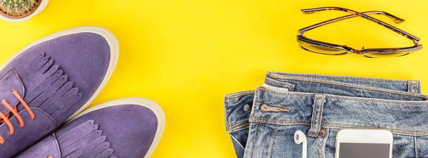 Flat lay of suede shoes, blue jeans, grey hat, cactus and smartphone headphones on bold yellow paper background with copy space. Overhead view of woman casual outfit. Long wide banner - Photo, Image