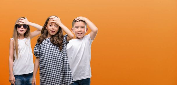 Группа мальчиков и девочек детей с оранжевым фоном напряжены с рукой на голове, шокированы стыдом и удивлением лица, сердитый и разочарованный. Страх и разочарование по ошибке
. - Фото, изображение