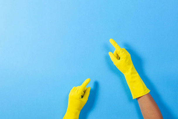 Woman hands with yellow rubber gloves points upwards with finger, over blue background - Foto, Imagen