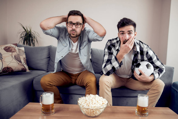 zwei männliche Freunde sehen Fußballspiel im Fernsehen, während sie Bier trinken und Popcorn essen. - Foto, Bild