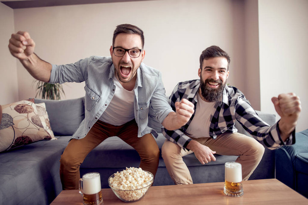 Friends celebrating a goal while watching a soccer game at home with popcorn and beer. - Foto, Imagem