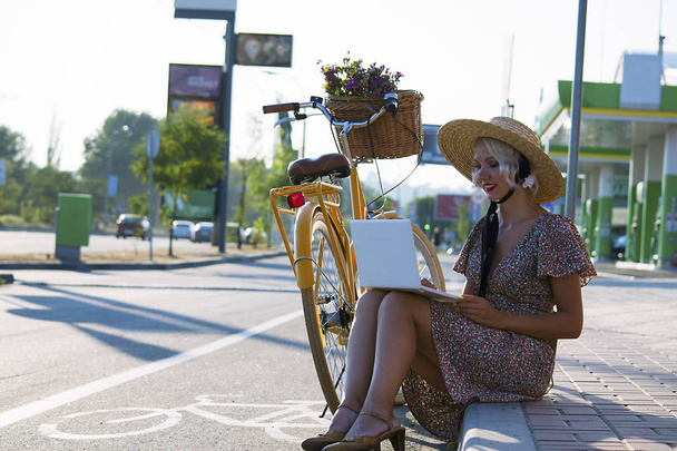 schattig meisje in een hoed kijkt laptop in een stad op de straat en een fiets - Foto, afbeelding