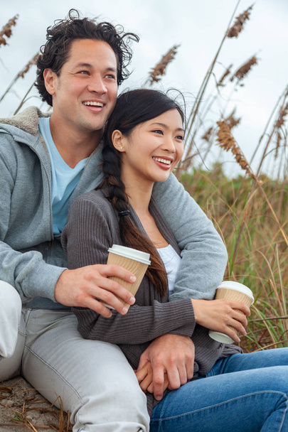 Young Asian Chinese man and woman, boy girl, couple sitting on a beach smiling with perfect teeth and drinking takeout cups of tea or coffee - Valokuva, kuva