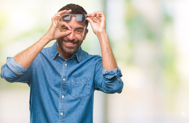 Adult hispanic man wearing sunglasses over isolated background doing ok gesture with hand smiling, eye looking through fingers with happy face. - 写真・画像