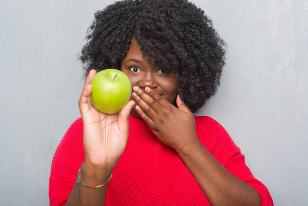 Joven mujer afroamericana sobre pared grunge gris comiendo boca cubierta de manzana verde con la mano conmocionada con vergüenza por error, expresión de miedo, asustada en silencio, concepto secreto
 - Foto, Imagen