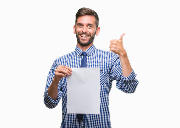 Young handsome man holding blank paper over isolated background happy with big smile doing ok sign, thumb up with fingers, excellent sign - Foto, immagini