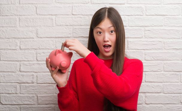 Young Chinese woman over brick wall holding piggy bank scared in shock with a surprise face, afraid and excited with fear expression - Photo, Image