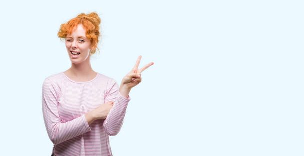 Young redhead woman smiling with happy face winking at the camera doing victory sign. Number two. - Photo, Image