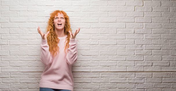 Young redhead woman standing over brick wall crazy and mad shouting and yelling with aggressive expression and arms raised. Frustration concept. - Photo, Image