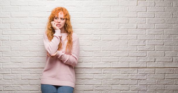 Mladá zrzka žena stojící nad cihlovou zeď stresovaní a nervózní s rukama na ústech kousat nehty. Úzkost problem. - Fotografie, Obrázek