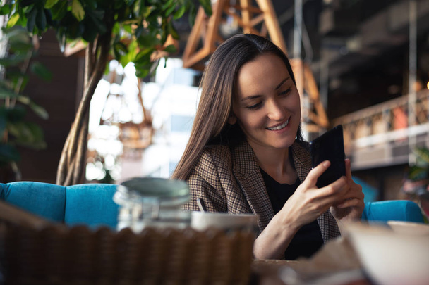 Young woman smiling with smartphone in hand at restaurant with blurred background. - Photo, Image