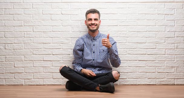 Молодий дорослий чоловік сидить над білою цегляною стіною, роблячи щасливі великі пальці вгору жестом рукою. Затвердження виразу, що дивиться на камеру з успіхом
. - Фото, зображення