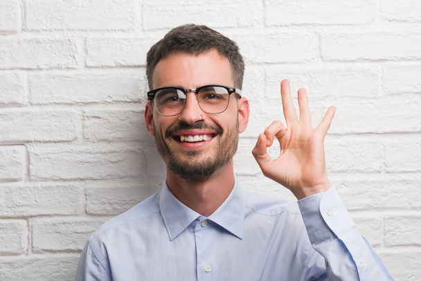 Молодой взрослый бизнесмен стоит над белой кирпичной стеной, делает хорошо знак с пальцами, отличный символ
 - Фото, изображение