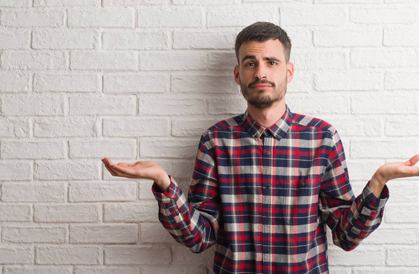 Young adult man standing over white brick wall clueless and confused expression with arms and hands raised. Doubt concept. - Photo, Image