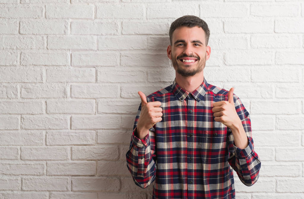 Mladý dospělý muž stojící nad bílá cihlová zeď úspěch znamení dělá pozitivní gesto rukou, palec se usmívá a šťastný. Při pohledu na fotoaparát s veselým výrazem, vítěz gesto. - Fotografie, Obrázek