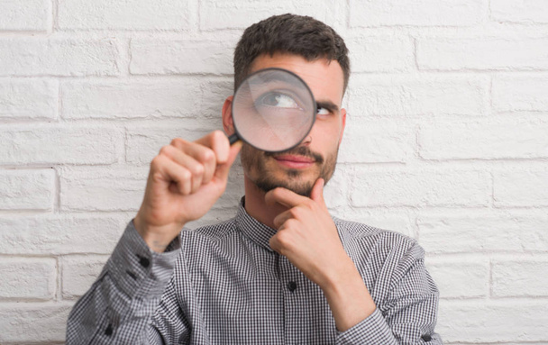 Young adult man over brick wall using magnifying glass serious face thinking about question, very confused idea - Photo, Image