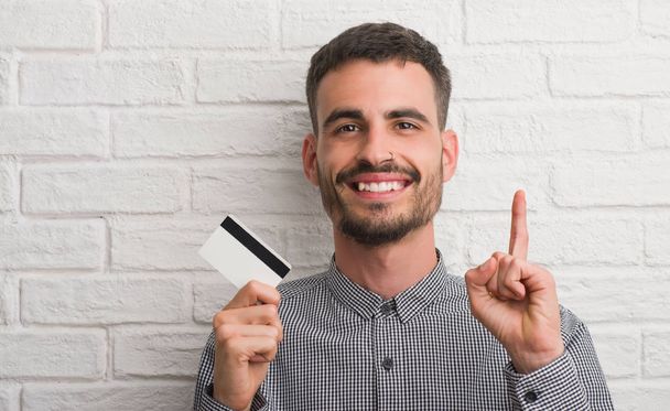 Fiatal felnőtt férfi-over tégla fal gazdaság hitelkártya meglepett egy ötlet, vagy ujját, boldog arc, az első kérdés - Fotó, kép