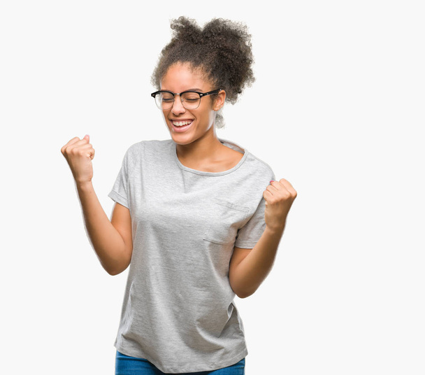 Młody afro american Kobieta w okularach na na białym tle bardzo szczęśliwy i podekscytowany robi z podniesionymi rękami, zwycięzca gest, uśmiechając się i krzyczy na sukces. Koncepcja celebracja. - Zdjęcie, obraz