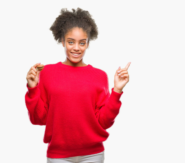 Młody afro american kobieta jedzenie czekolada makaronik na białym tle bardzo szczęśliwy, wskazując z dłoni i palców z boku - Zdjęcie, obraz