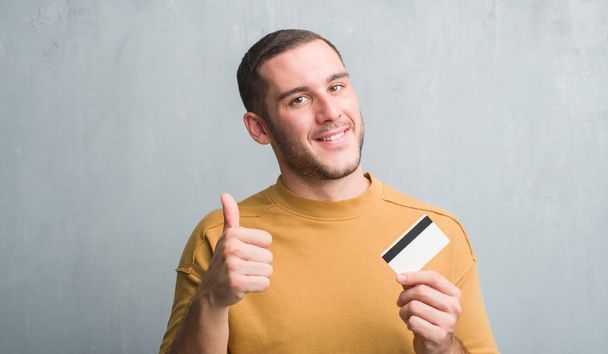 Homem caucasiano jovem sobre parede grunge cinza segurando cartão de crédito feliz com grande sorriso fazendo sinal ok, polegar para cima com os dedos, excelente sinal
 - Foto, Imagem