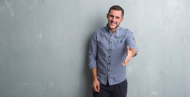 Молодий кавказький чоловік над сірою гранжевою стіною посміхається доброзичливо пропонуючи рукостискання як привітання і вітання. Успішний бізнес
. - Фото, зображення