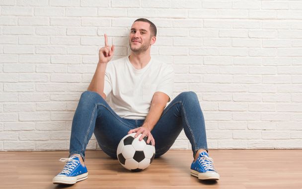 Young caucasian man sitting over white brick wall holding soccer football ball surprised with an idea or question pointing finger with happy face, number one - Photo, Image