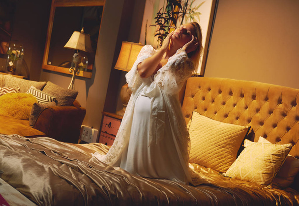 Young relaxed attractive blond pregnant woman wearing white rest gown posing on the bed in fashionable luxury rich bedroom at home alone. Romantic mood, modern cozy, interior. Brown, beige yellow colour - Foto, afbeelding