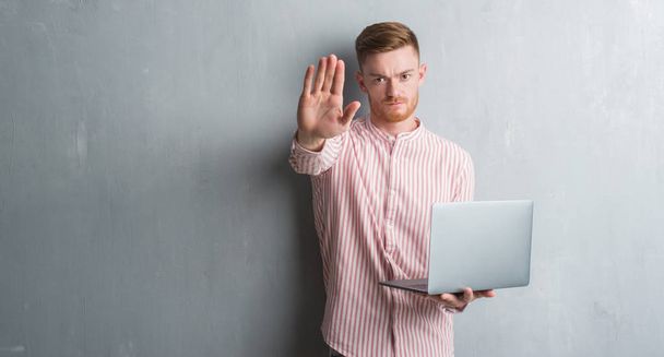 Genç Kızıl saçlı adam tutarak ve açık eliyle dur işareti ile ciddi ve kendine güvenen ifade, savunma hareketi yaparken bilgisayar dizüstü kullanarak gri grunge duvarın üzerinden - Fotoğraf, Görsel