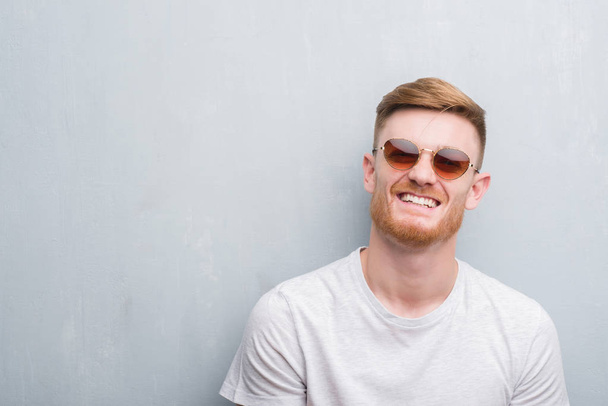 Genç Kızıl saçlı adam ayakta ve diş gösterilen kendine güvenen bir gülümsemeyle gülümseyerek mutlu bir yüz ile retro güneş gözlüğü takıyor gri grunge duvarın üzerinden - Fotoğraf, Görsel