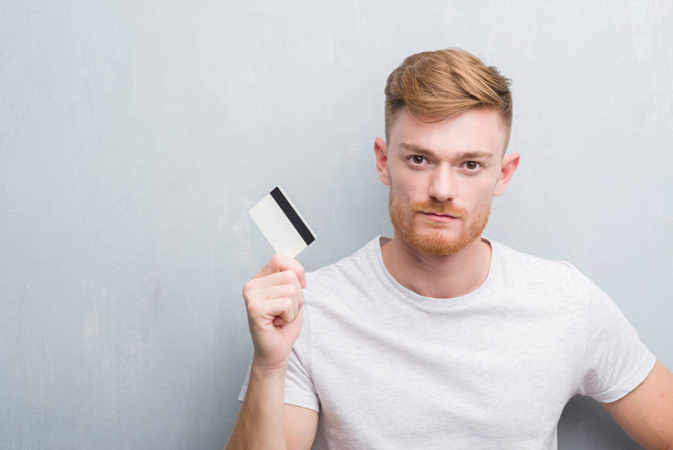 junger rothaariger Mann über grauer Grunge-Wand mit Kreditkarte und selbstbewusstem Gesichtsausdruck - Foto, Bild