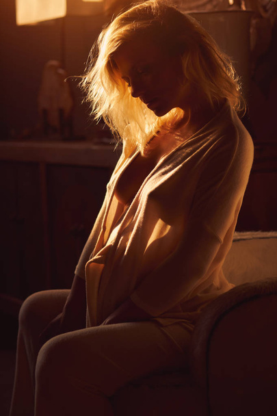 Sensual beautiful thoughtful pensive pregnant woman sitting in bedroom at home. Relaxed blond fashionable young female in morning gown or bathrobe sitting with naked breast. Shadows, romantic mood - Photo, image