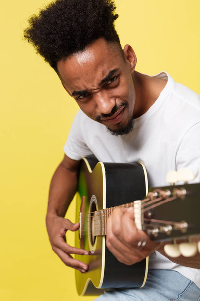 Młody przystojny African American retro stylu gitarzysta gra gitara akustyczna na żółtym tle złotym tle. - Zdjęcie, obraz